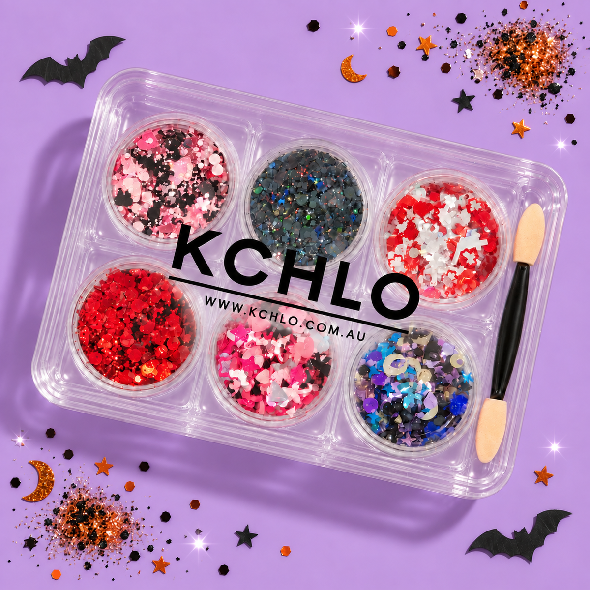 A clear plastic makeup glitter tray with six round compartments filled with assorted Halloween-themed glitter in red, pink, black, blue, and silver. A dual-ended sponge applicator rests on the right side of the tray.